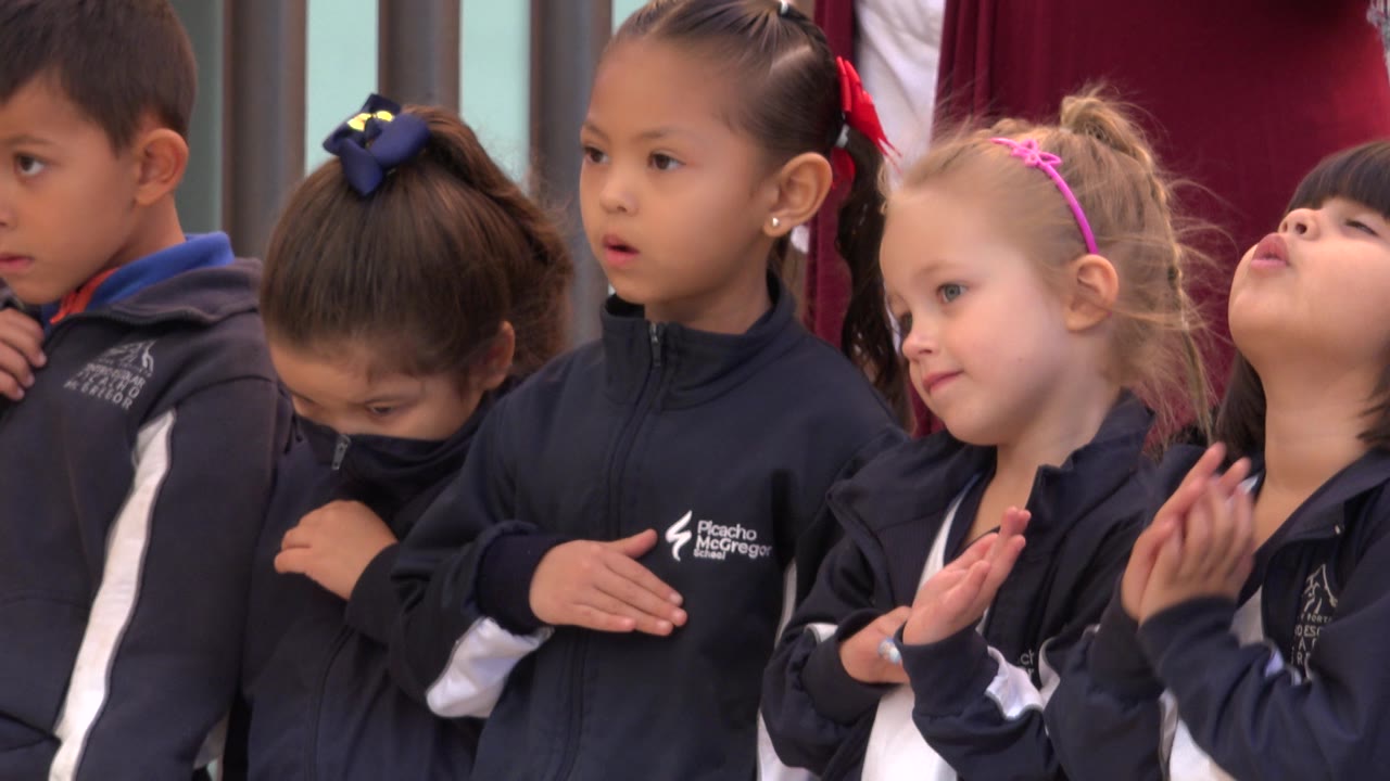 A close group of children in matching dark jackets stands in San Jose del Cabo, Mexico, as a girl with a pink headband lifts her hands and another child tilts her head back.