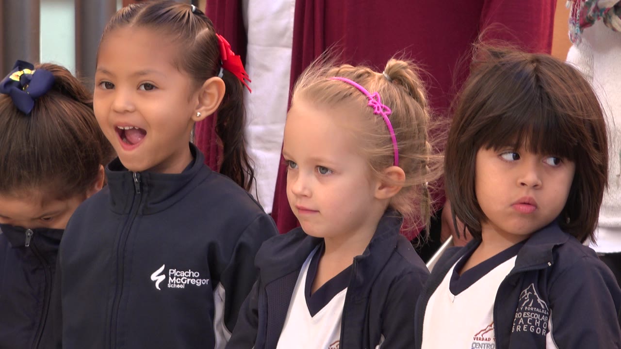 Four school-age girls stand shoulder to shoulder in San Jose del Cabo, Mexico, with dark jackets, light shirts, hair bows, and red fabric behind them.