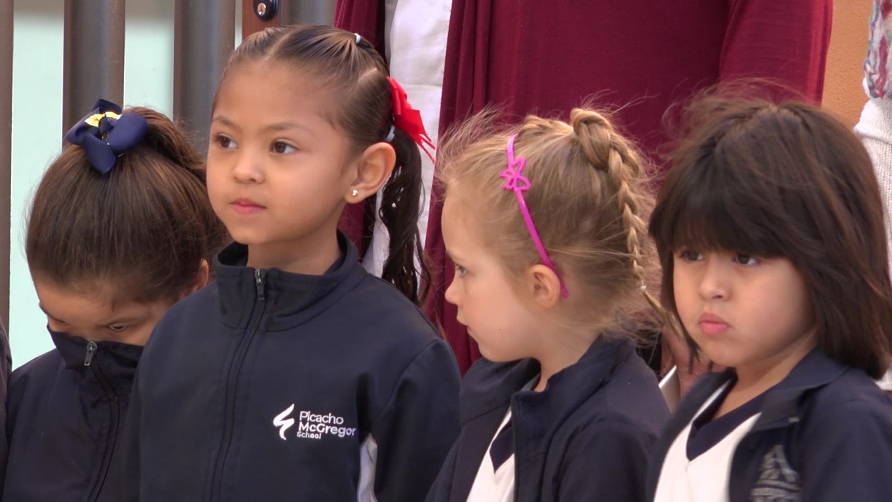 Four young girls standing close together outdoors with adults behind them.