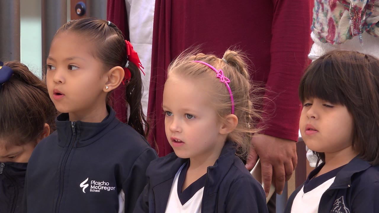At 0:02, three young girls are most prominent in the frame, including a girl with a red hair ribbon, a blonde girl with a pink headband, and a dark-haired girl in navy and white clothing.