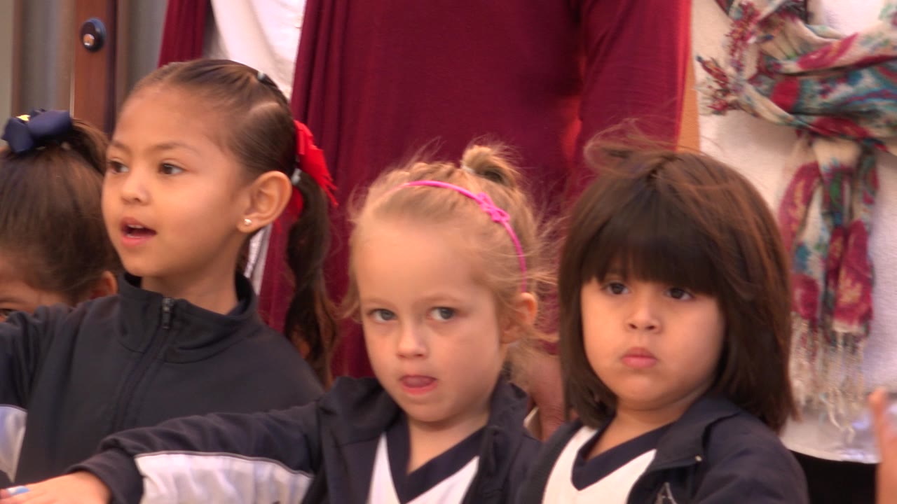 At 0:00, young children in dark uniforms stand in a close group in San Jose del Cabo, Mexico, with cropped adults behind them and one child looking off frame.