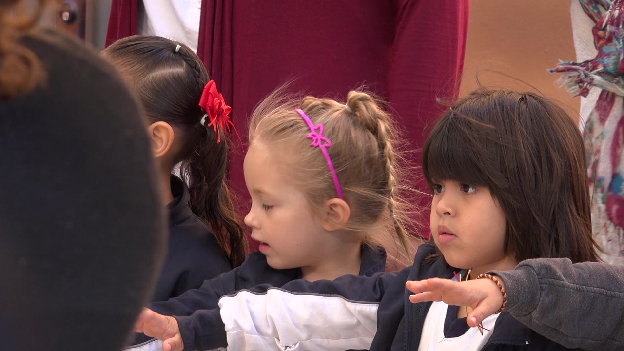 At 0:00 in San Jose del Cabo, young children stand shoulder to shoulder with one dark-haired child looking ahead while adults fill the background.