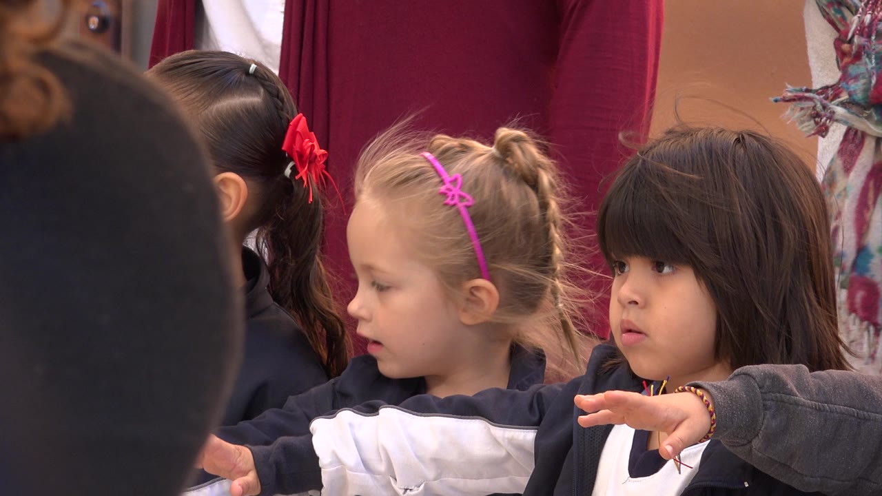 At 0:01, a blonde child with a pink headband looks left beside a dark-haired child extending her arm, with adult clothing visible behind them in San Jose del Cabo.