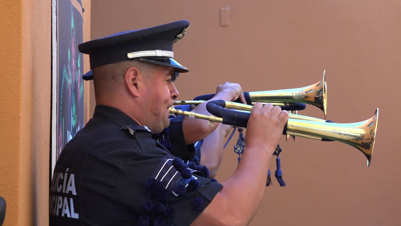 Uniformed police officer playing two brass trumpets beside a peach wall.