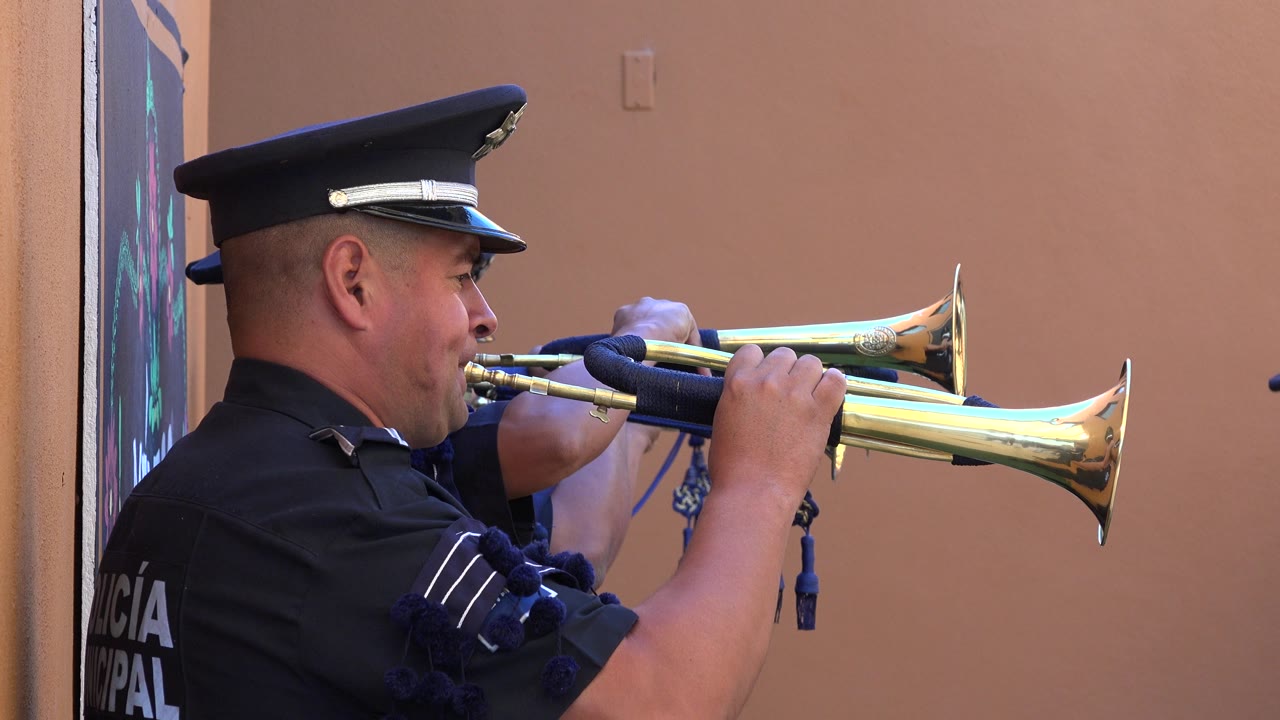 A uniformed man in San Jose Del Cabo, Mexico plays a brass trumpet in side profile beside a peach-colored wall at the start of the clip.