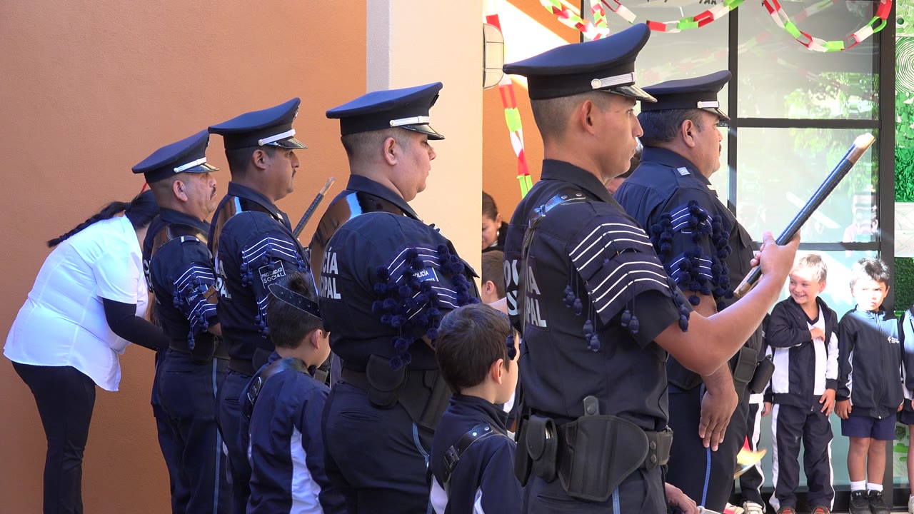 Uniformed police officers stand in profile at a San Jose del Cabo ceremony while children in navy uniforms gather near a doorway behind them.