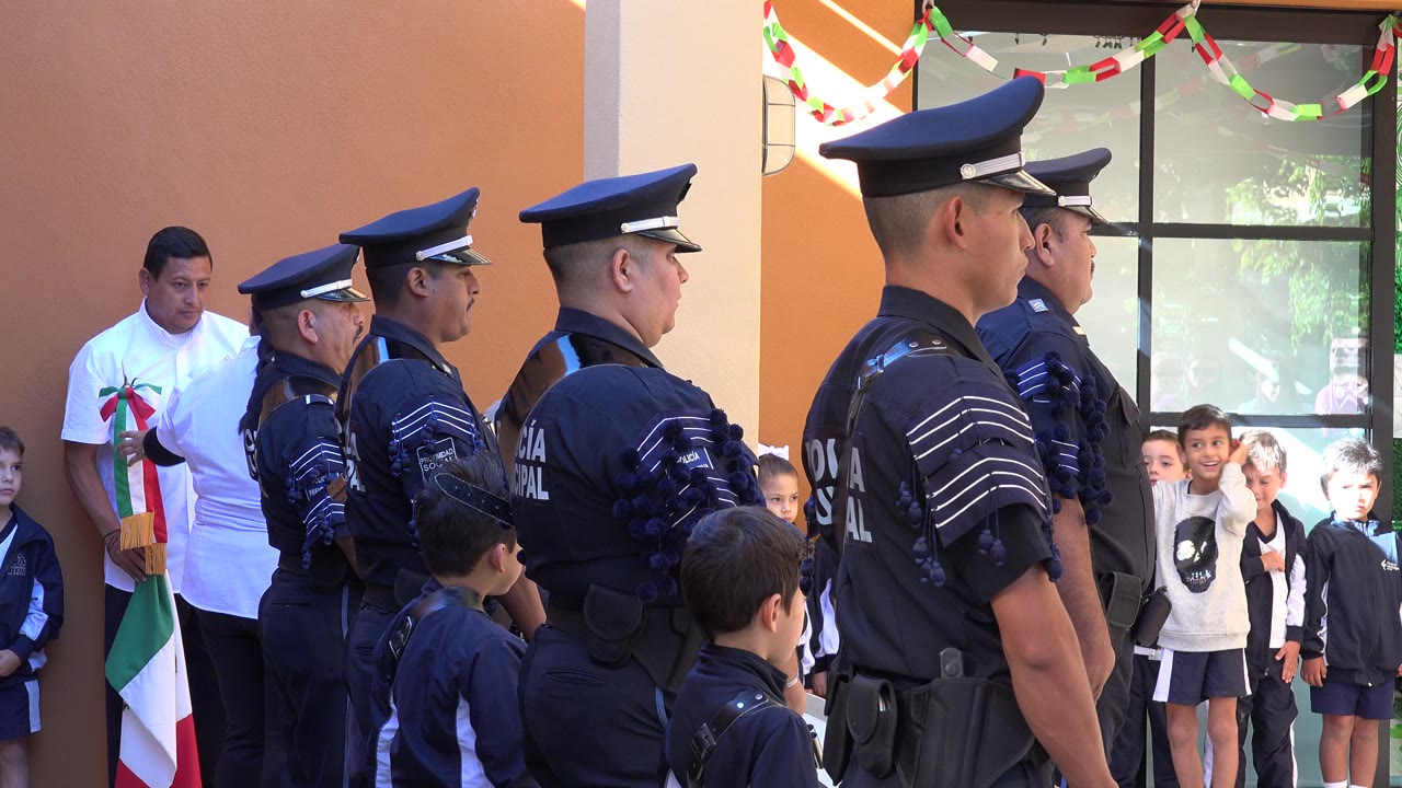 Uniformed police officers stand in profile outside a school in San Jose del Cabo, Mexico, with children and an adult visible near the orange wall and window.