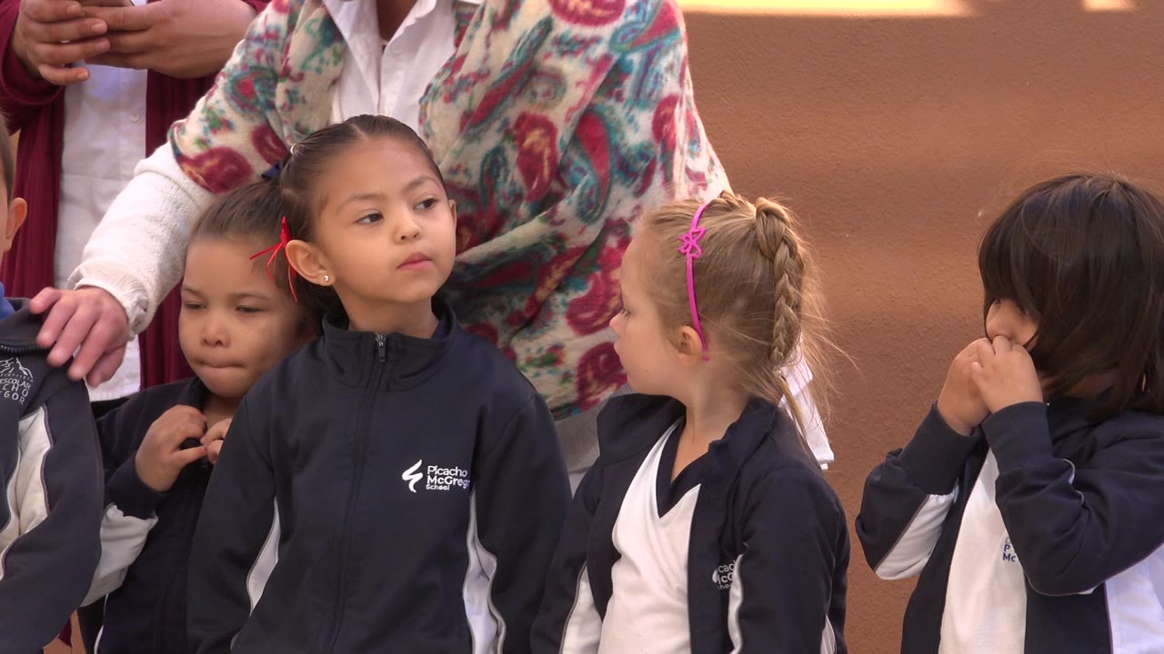 Young girls in navy and white team warmups stand closely together in San Jose del Cabo, Mexico, with cropped adults behind them on a reddish outdoor court.