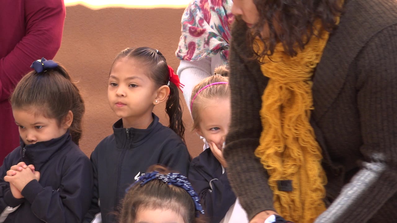 Children in San Jose Del Cabo, Mexico stand in a tight group as an adult in a dark sweater and yellow scarf leans over them.