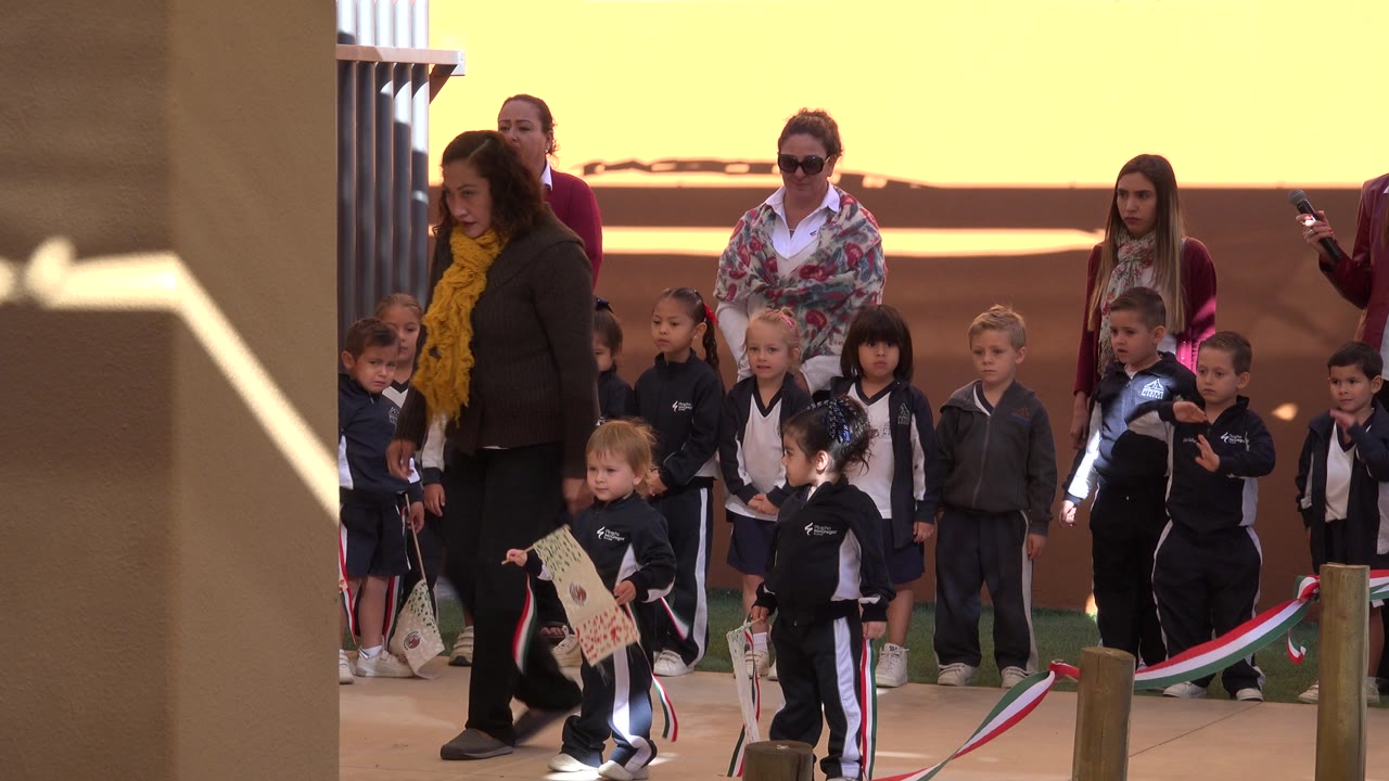 Young children in dark uniforms stand with adult women during a San Jose del Cabo parade or ceremony, with tan columns and a bright yellow wall behind them.
