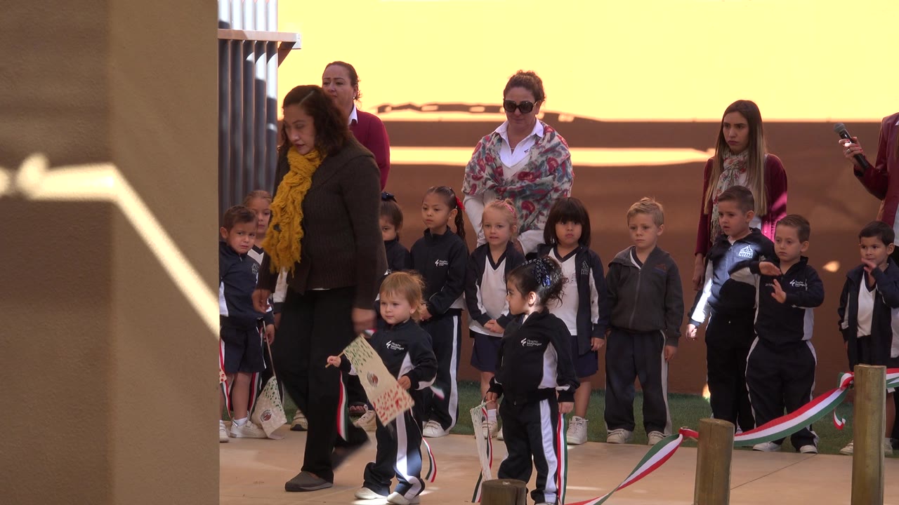A line of children in dark school-style uniforms waits in the midground in San Jose del Cabo, with adult women behind them and a child holding a decorated flag in front.