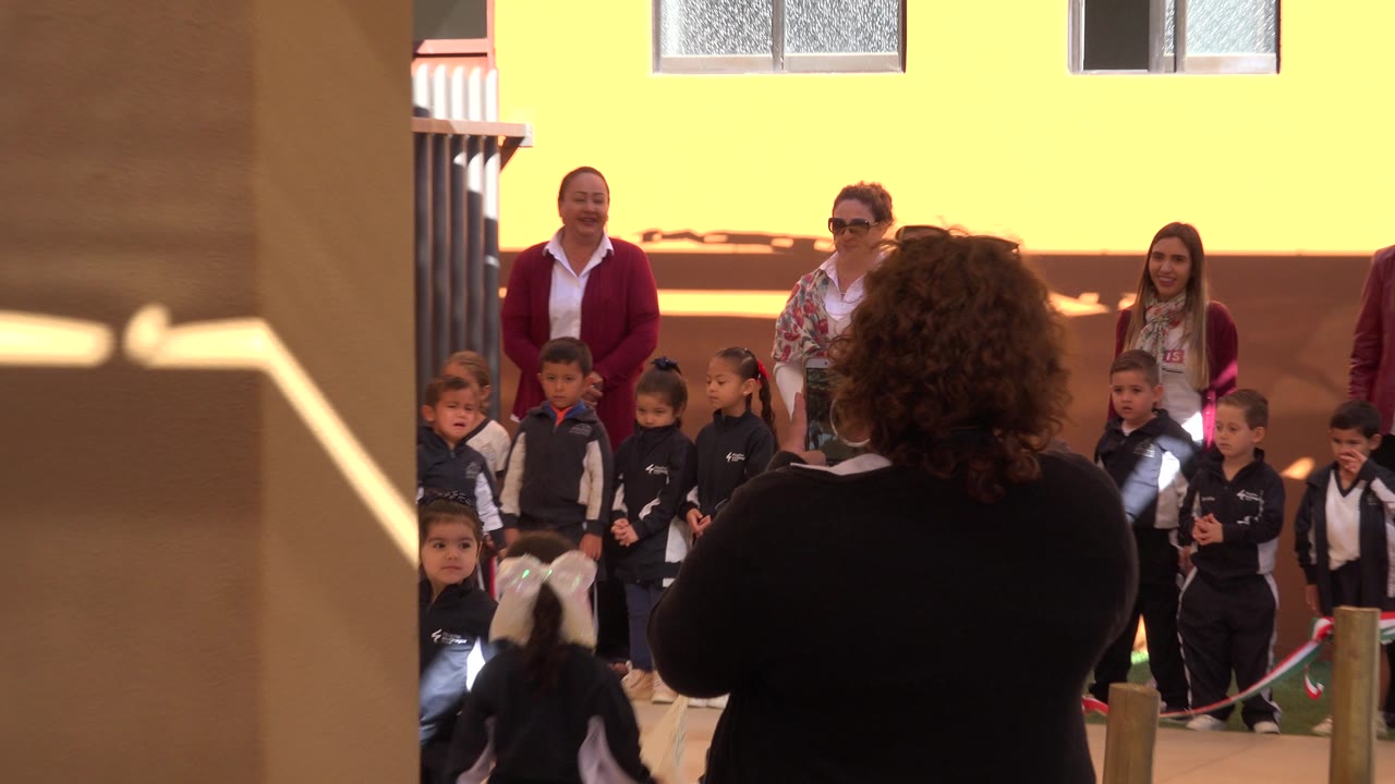 Children in school uniforms stand with adults in a sunny courtyard.