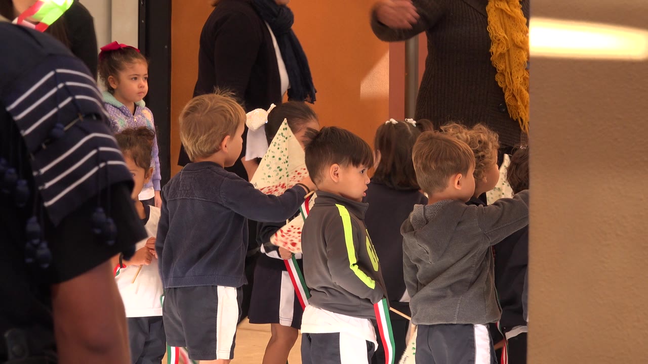 Children in school uniforms holding small flags near an orange wall.