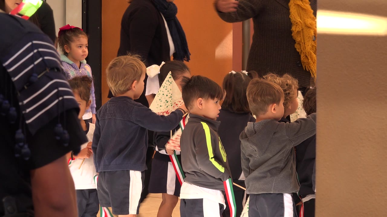 A blond child faces sideways among classmates in San Jose del Cabo, Mexico, while children around him hold small decorated flags and red, white, and green ribbons.