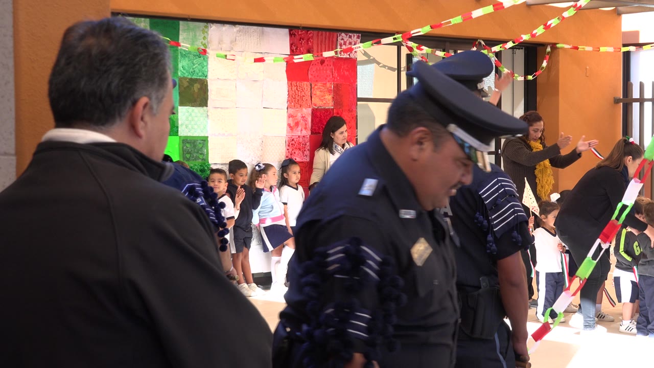A uniformed police officer is close to camera as children stand in rows behind him in the decorated San Jose del Cabo community space.