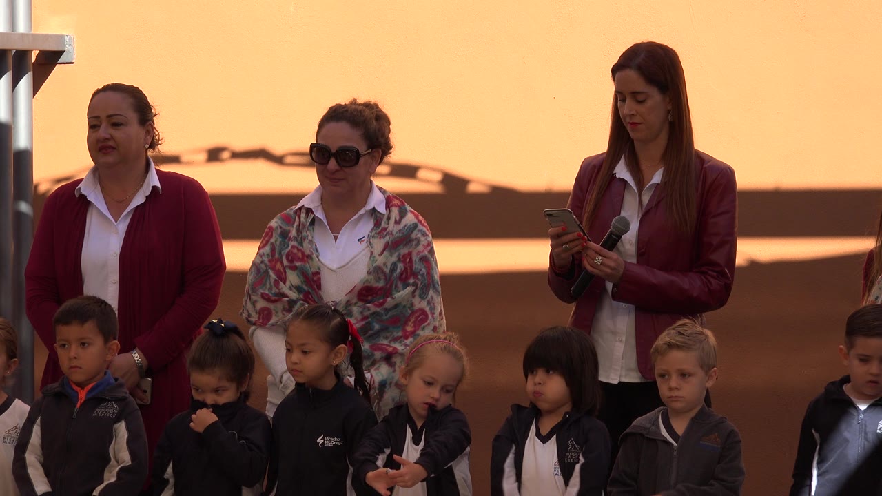 A row of young soccer players stands in the foreground in San Jose del Cabo while three adult women remain behind them, including a presenter looking down at a phone.
