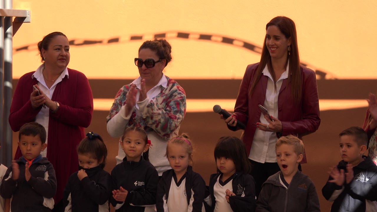 In San Jose del Cabo, Mexico, young children stand in front while three adult women behind them clap during a school event under warm orange light.