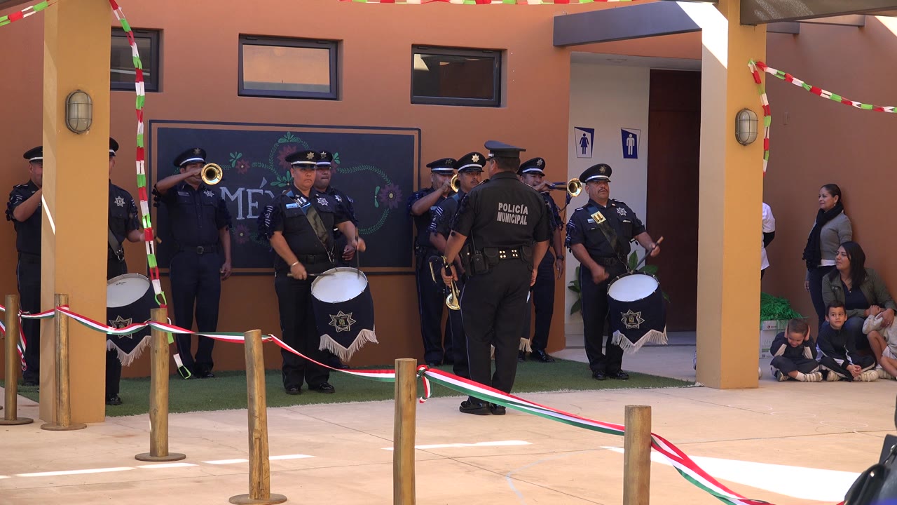A drummer stands near center as other uniformed officers hold brass instruments, with seated children and adult onlookers visible along the right side of the San Jose del Cabo courtyard.