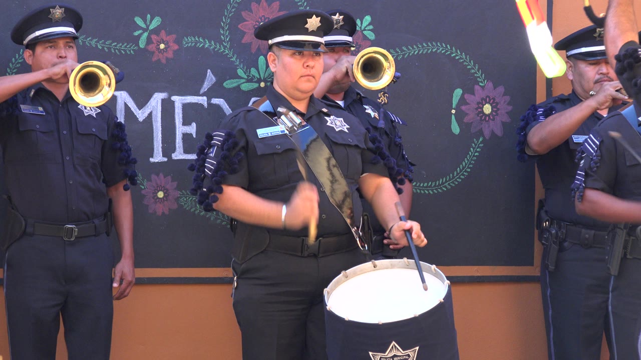 At 0:01 in San Jose Del Cabo, Mexico, the foreground drummer raises one stick as a trumpet bell points toward the camera from behind her shoulder.