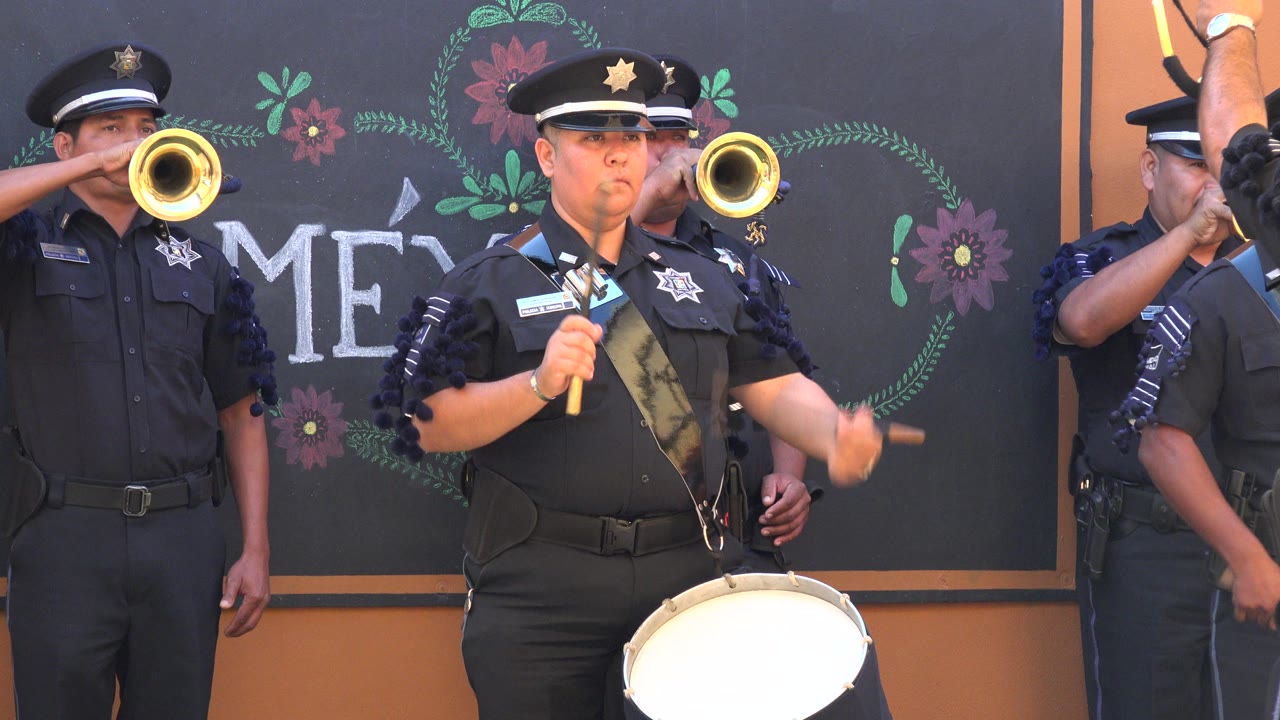 A uniformed drummer in San Jose del Cabo, Mexico raises both drumsticks as two trumpet bells appear behind him against the decorated dark backdrop.