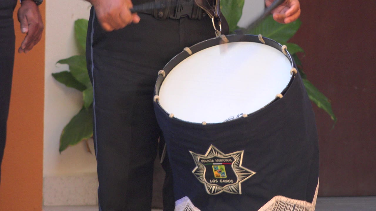 At 0:00 in San Jose del Cabo, a black fabric-covered drum with a white head hangs from a strap in front of a uniformed performer's torso.