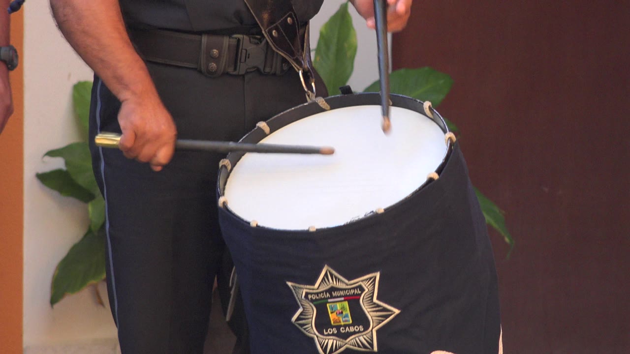 Two drumsticks cross over the white drumhead while a person in black pants holds the instrument in San Jose del Cabo, Mexico, with the emblem partly centered on the drum cover.