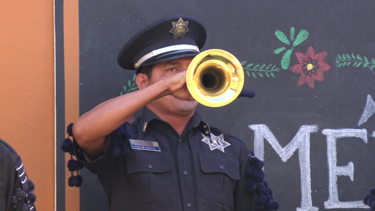 The brass horn bell sits near the center of the frame as the uniformed musician continues playing against the chalkboard-style background in San Jose del Cabo, Mexico.