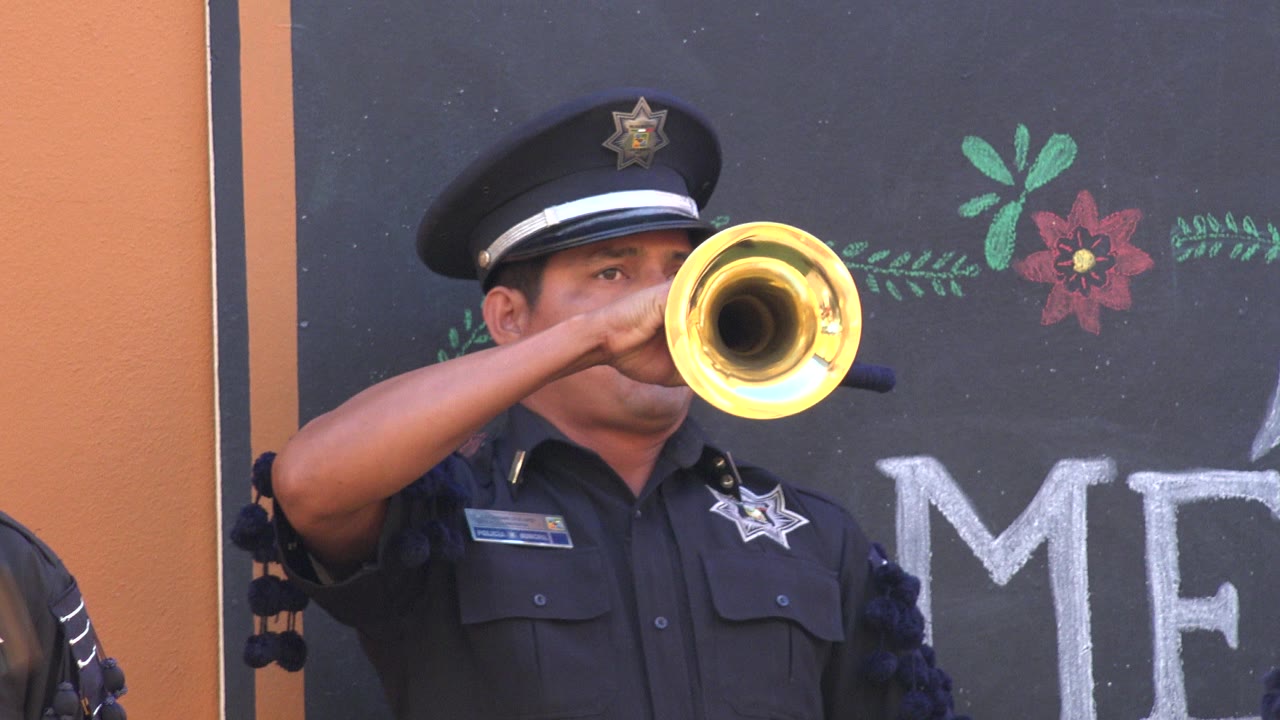 At 0:01, the San Jose del Cabo officer remains framed chest-up as the trumpet bell faces the camera and chalk flowers sit above the partial lettering behind him.
