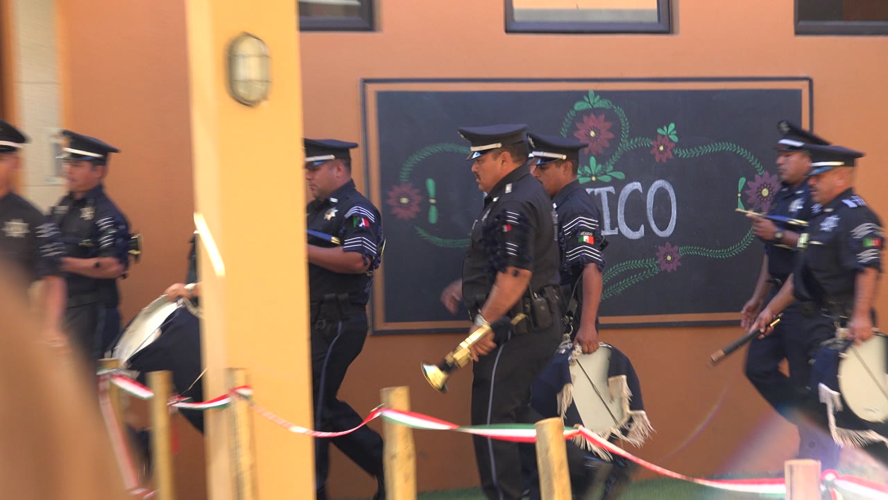 Uniformed policemen march in San Jose Del Cabo, Mexico, beside an orange wall with a chalkboard-style Mexico sign while blurred spectators frame the foreground.