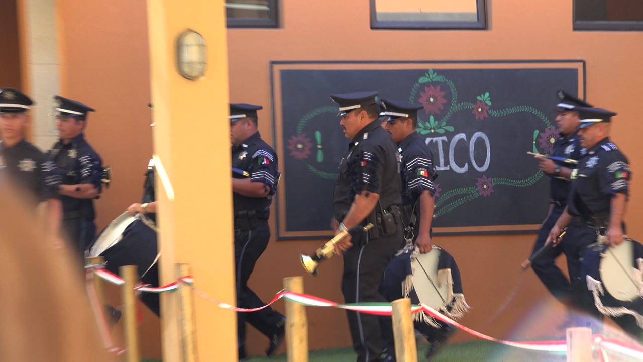 Police musicians in dark uniforms continue through the San Jose Del Cabo courtyard, with a drum, brass instrument, rope barrier, and the Mexico sign behind them.