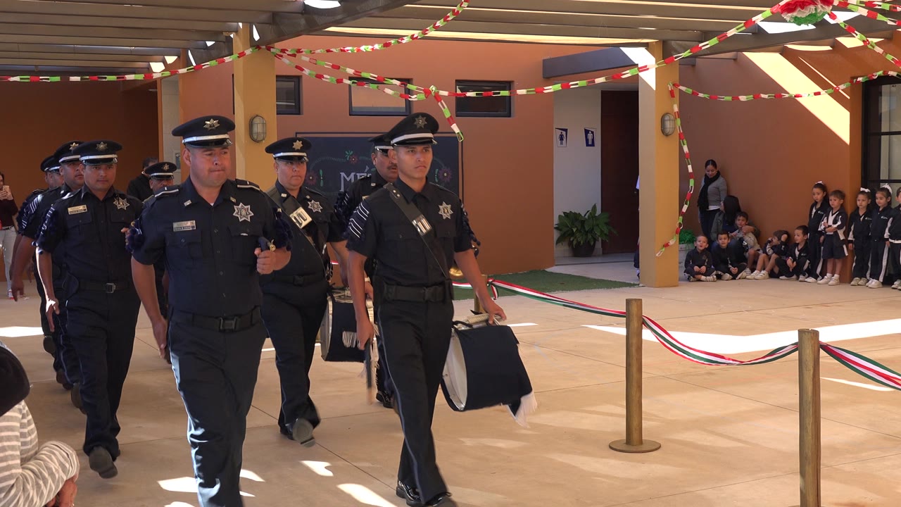 Uniformed police officers march toward the camera in San Jose del Cabo, Mexico, under red, white, and green streamers with seated children visible along the wall.
