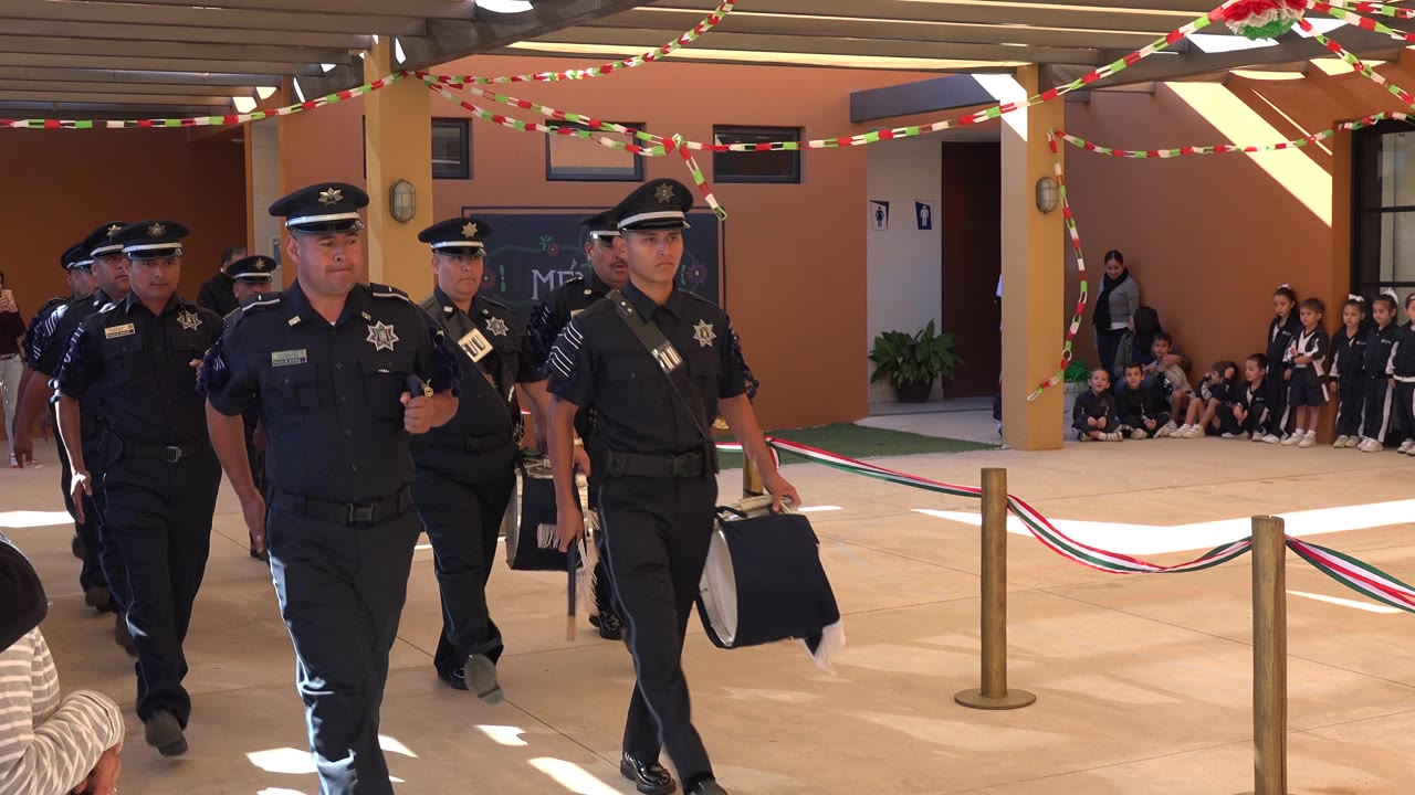 A line of police officers in dark uniforms advances through the San Jose del Cabo covered space as children sit in the background near wall signs.