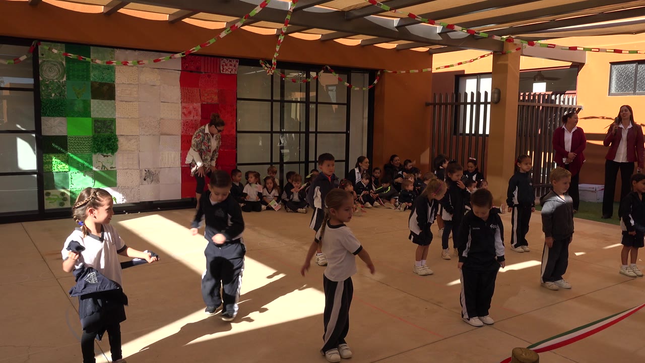 Several children shift positions in the San Jose del Cabo school courtyard as two adults stand near the back and a seated group watches by the wall.