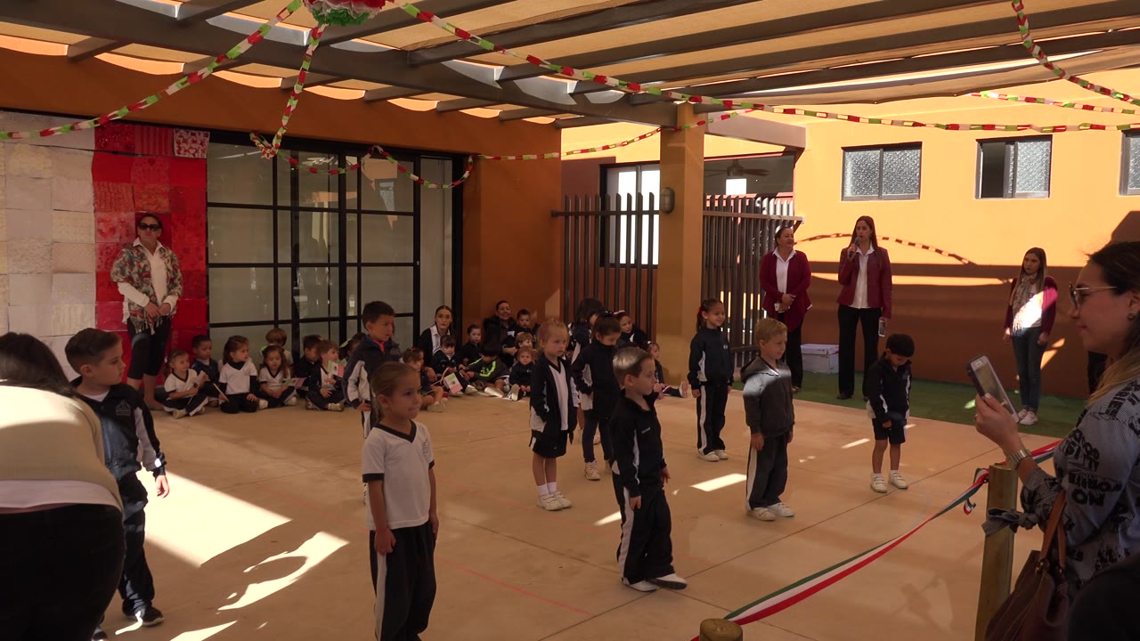 Children in uniforms stand spaced across a covered courtyard in San Jose del Cabo, Mexico, with seated children along the wall and adults watching from the back.