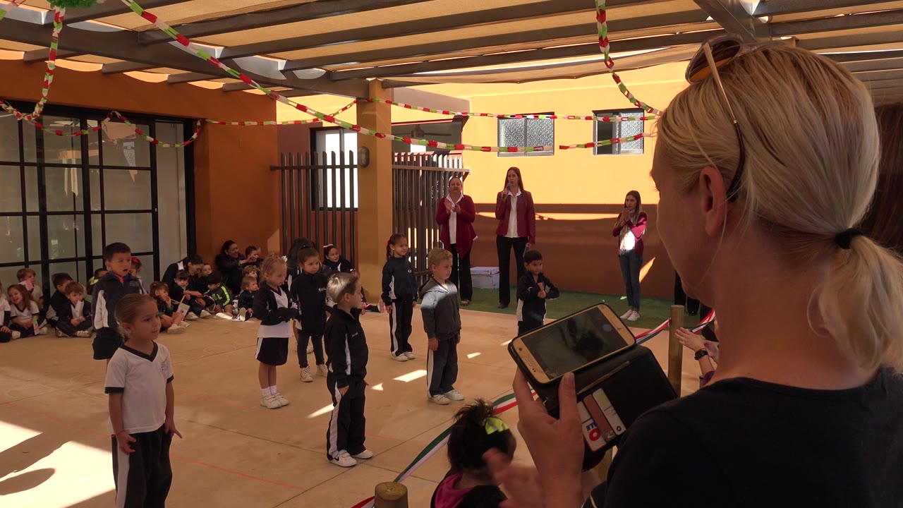 Children in track suits stand on a covered patio as adults and seated classmates watch.