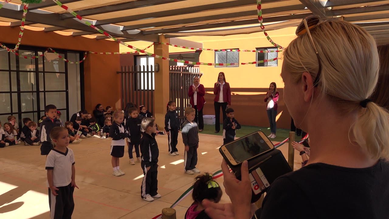 At 0:01 in San Jose del Cabo, Mexico, the foreground woman with a phone frames the children in navy and white uniforms as adults stand near the back wall.