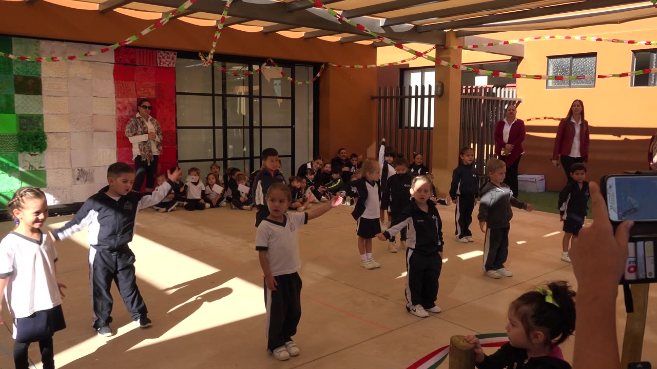 Children in school uniforms stand in a decorated courtyard while classmates sit along the wall.