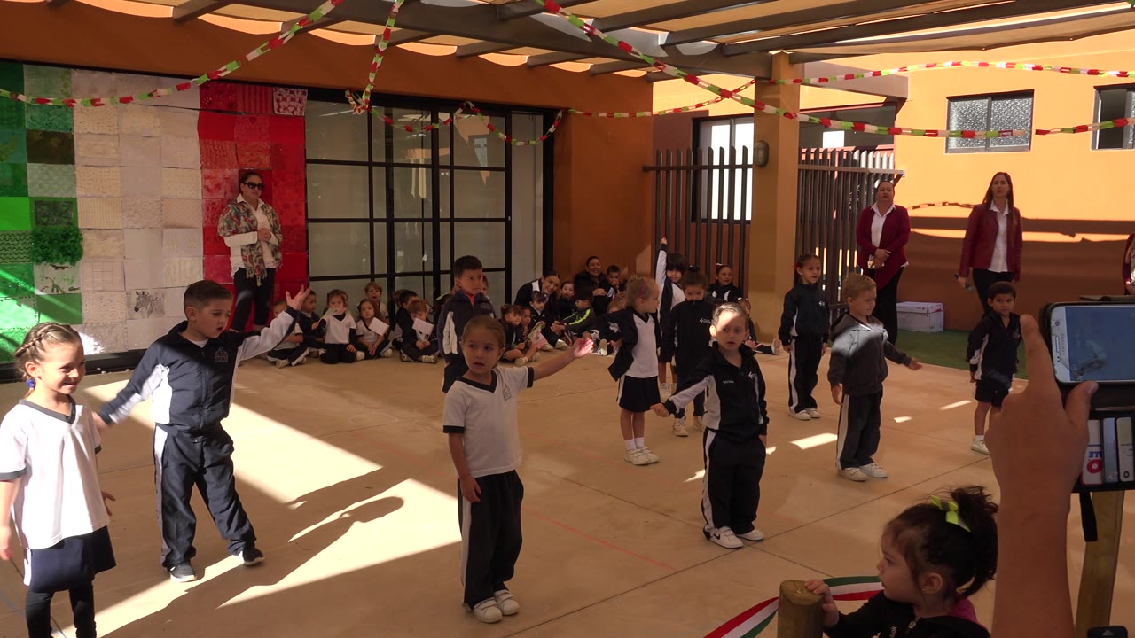 Children in uniforms stand and watch in the San Jose del Cabo courtyard while a foreground hand holds a smartphone, with classmates seated against the back wall.