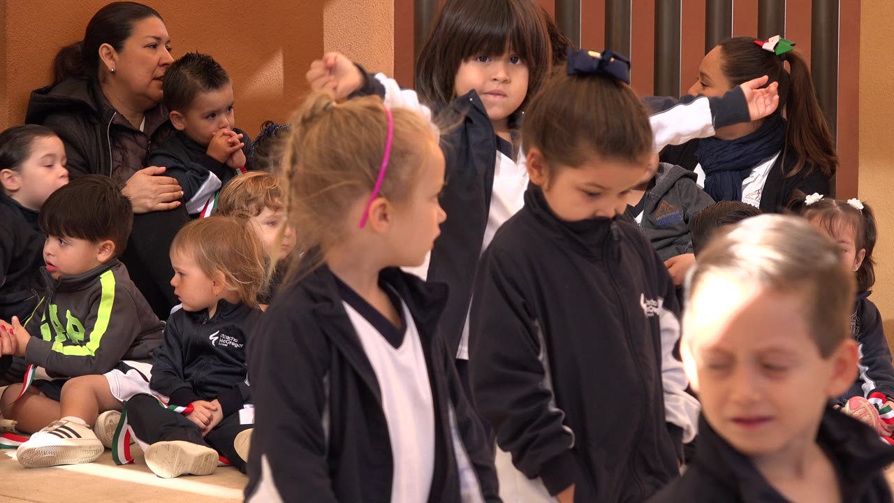 Two girls in navy jackets stand near the foreground as seated children and adults wait behind them in the San Jose del Cabo practice space.