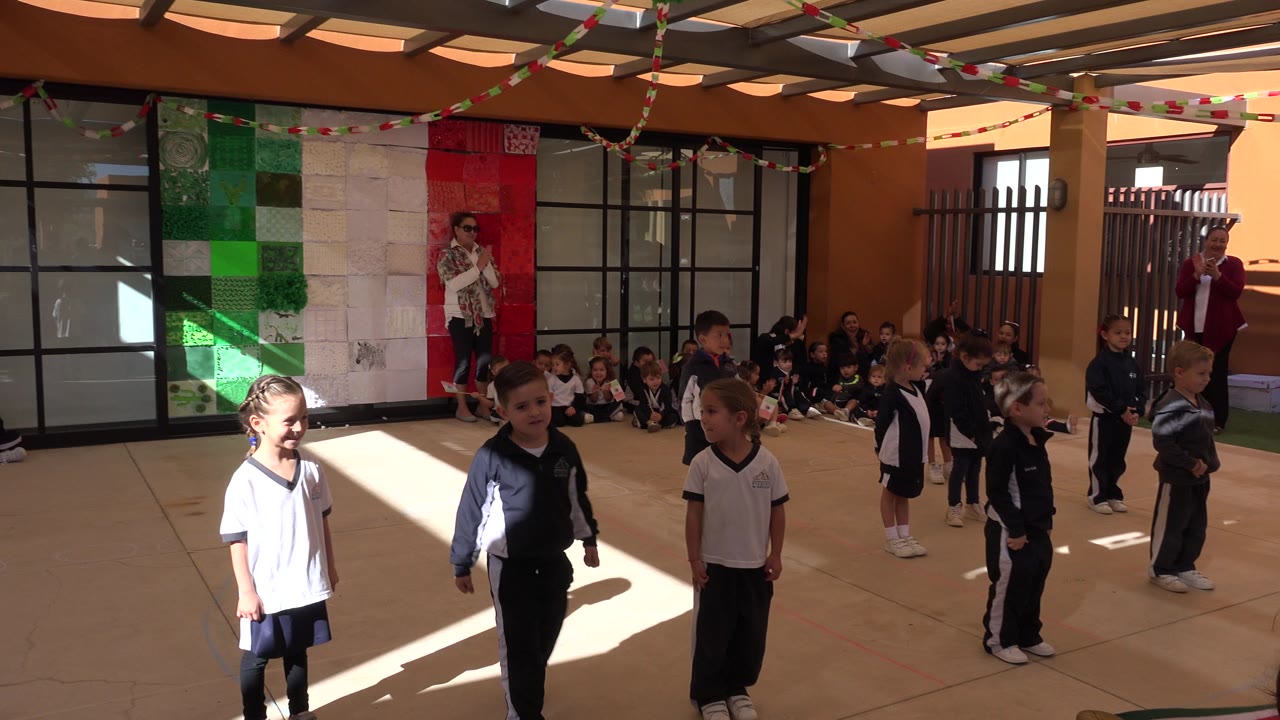 Children in school uniforms stand on a sunlit classroom floor while classmates sit behind them.