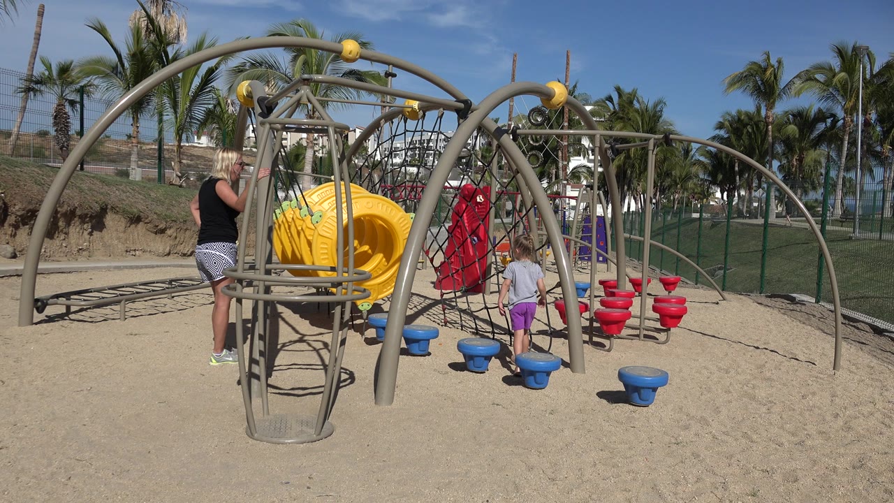 Woman watching children at a sandy playground with a yellow tunnel.
