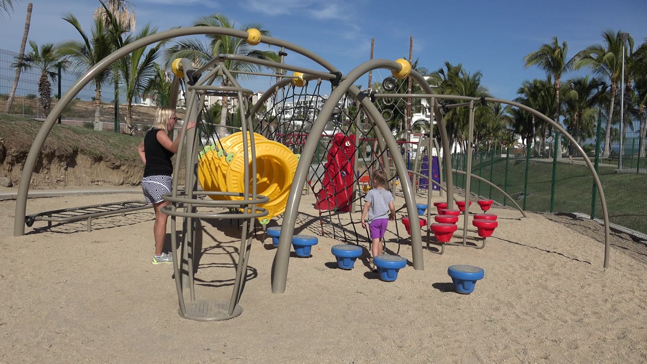 At 0:01, the children remain near the yellow tunnel and blue stepping pods while the woman watches from the left side of the sandy playground in San Jose Del Cabo, Mexico.