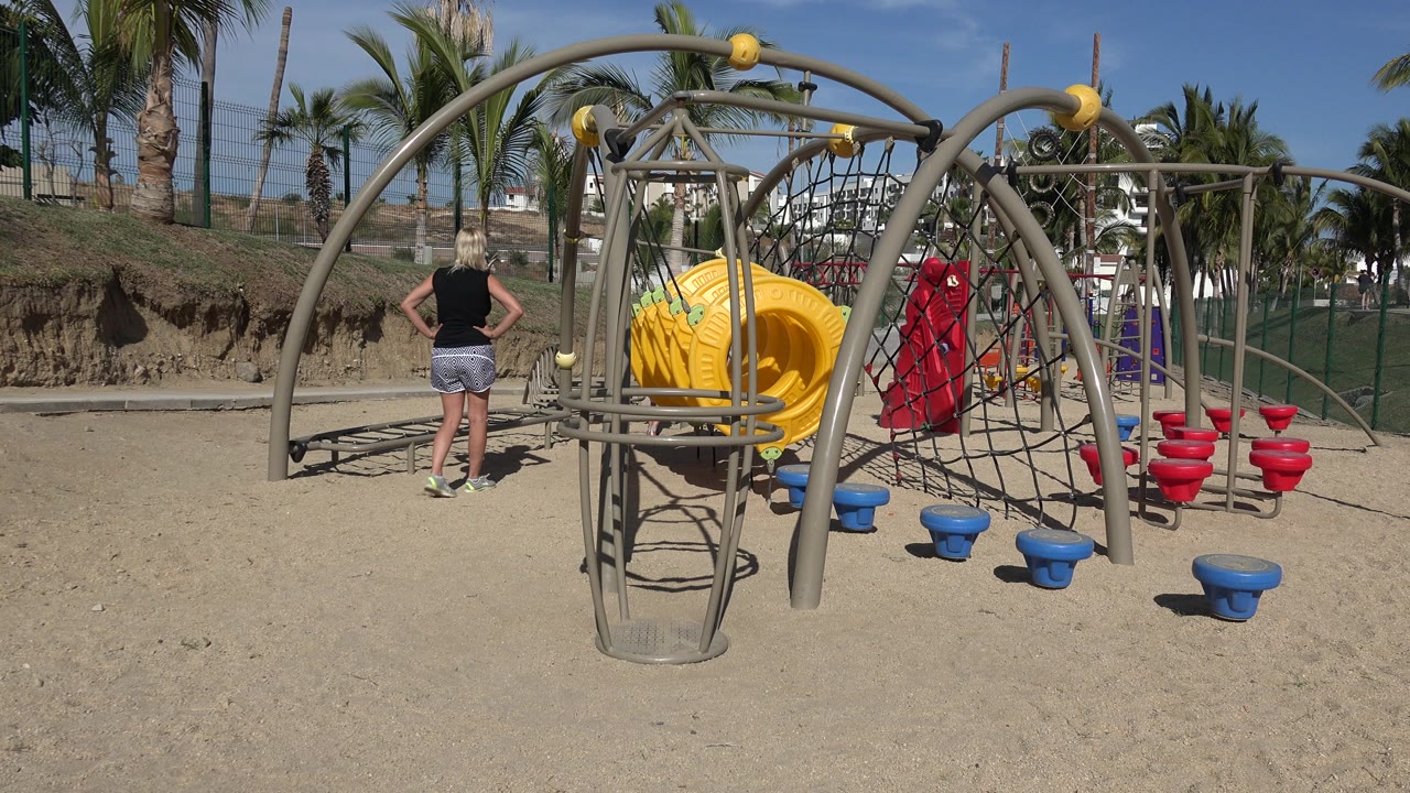 Sandy playground with climbing frame, slide, and adult viewed from behind.