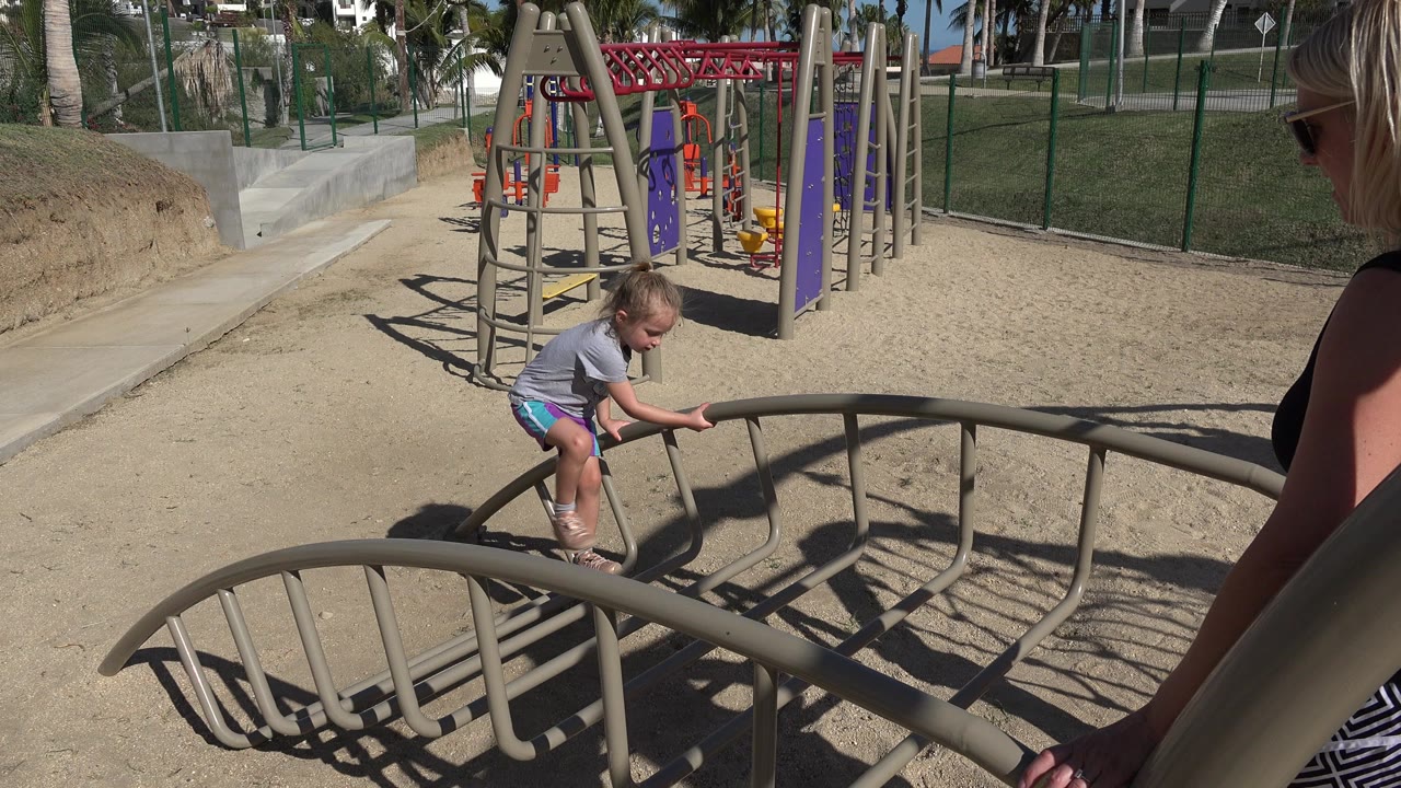Child stepping onto a curved metal playground frame while an adult watches nearby.