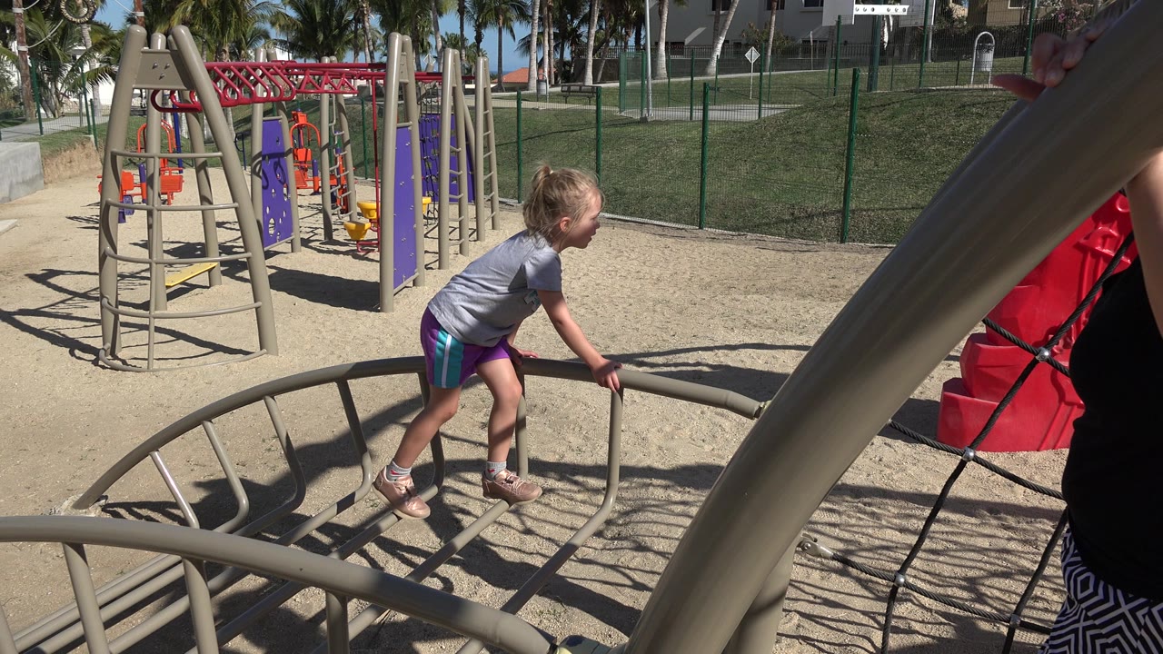A young girl climbs onto a beige metal playground structure in San Jose del Cabo, Mexico, with red and purple play equipment, fencing, grass, and palm trees in the background.