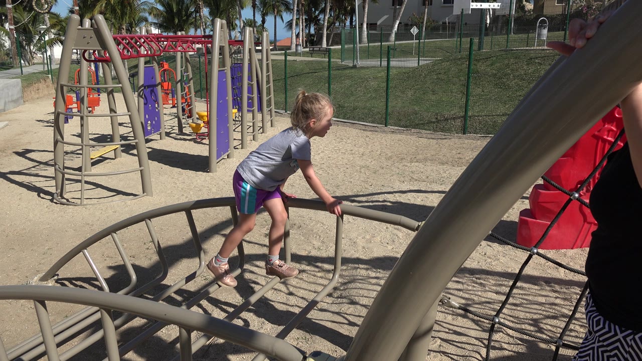 The girl grips a curved beige playground bar in San Jose del Cabo, Mexico, while part of an adult woman appears at the right edge near a red climbing panel.