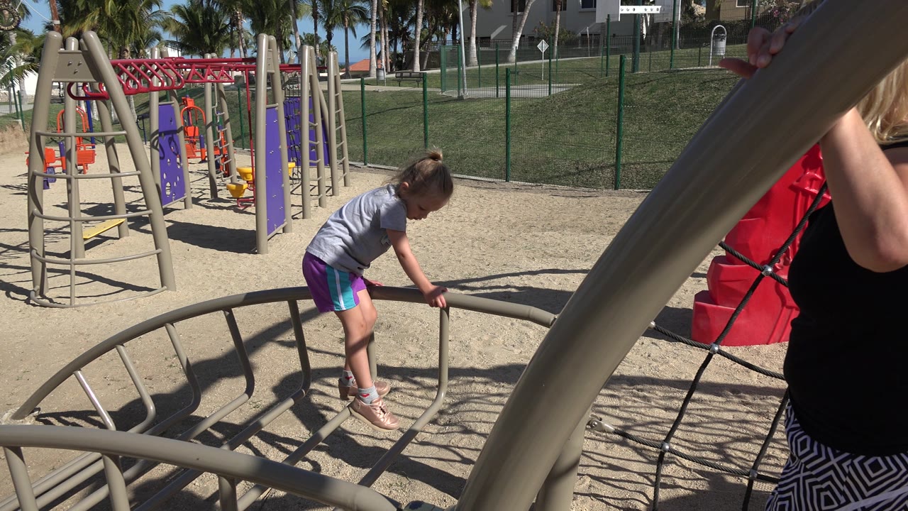 Girl bends over a playground rail with a woman partly visible beside the climbing net.