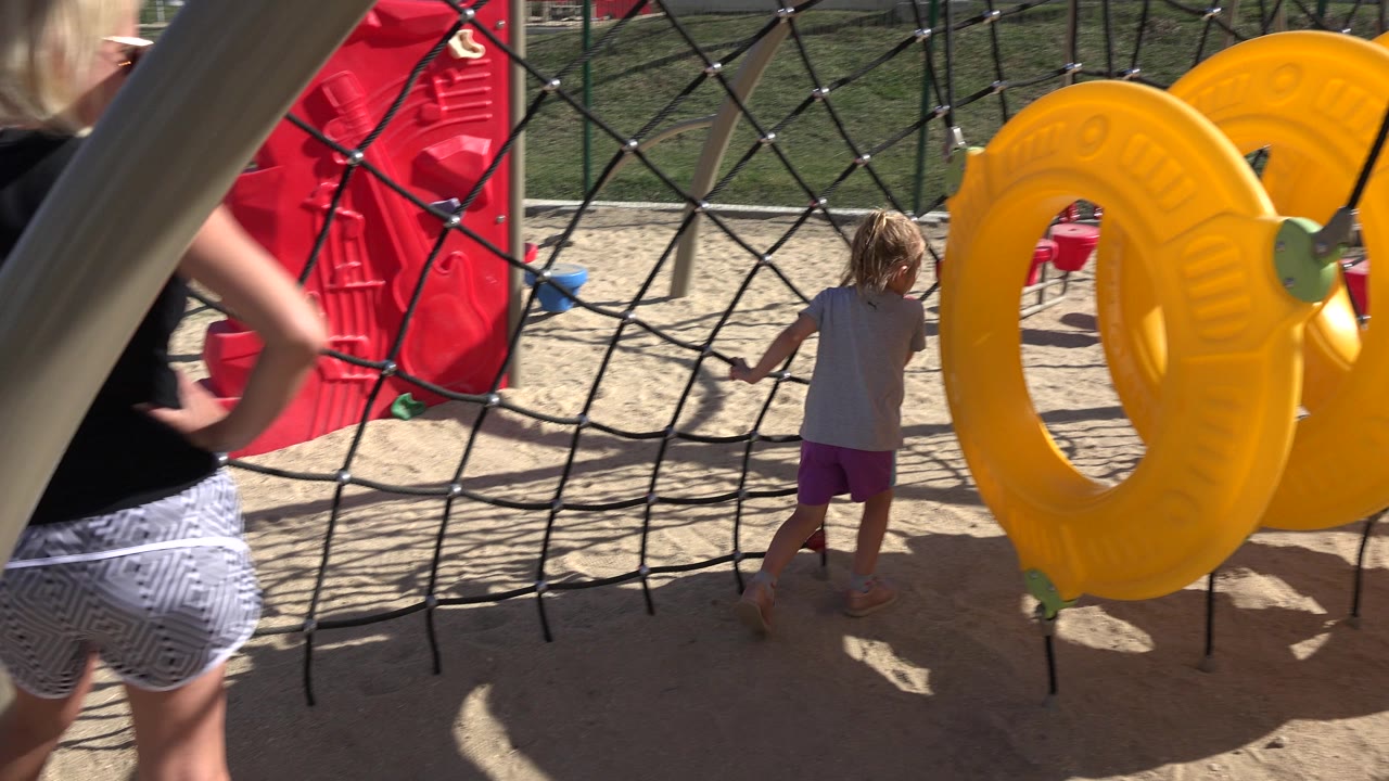 At 0:00 in San Jose Del Cabo, a child stands by a black rope climbing net while large yellow ring panels fill the right side and an adult is partly visible at the left edge.