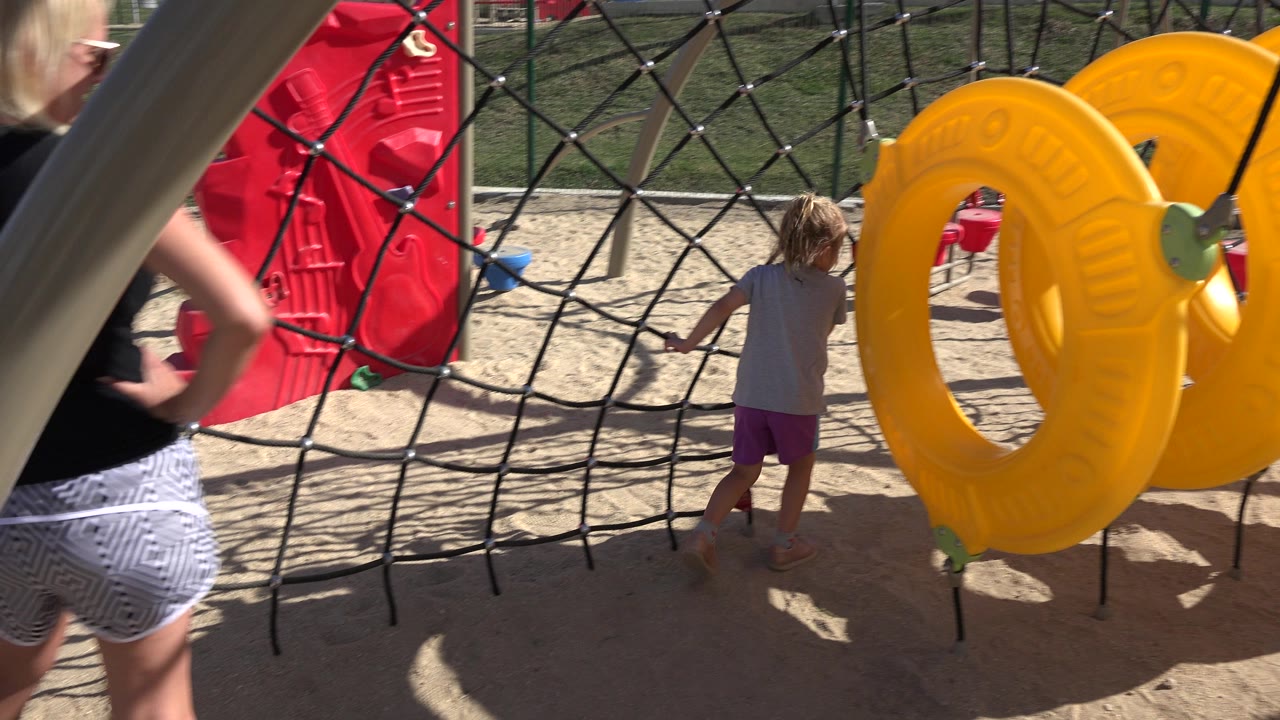 At 0:01 in San Jose Del Cabo, the child faces the rope net on sandy ground as yellow hanging rings and a red playground panel frame the play area.