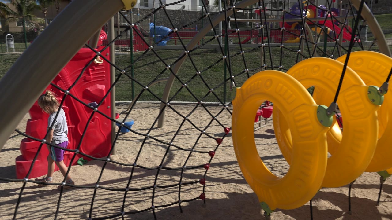 Child near red playground structure behind rope netting and yellow play seats.